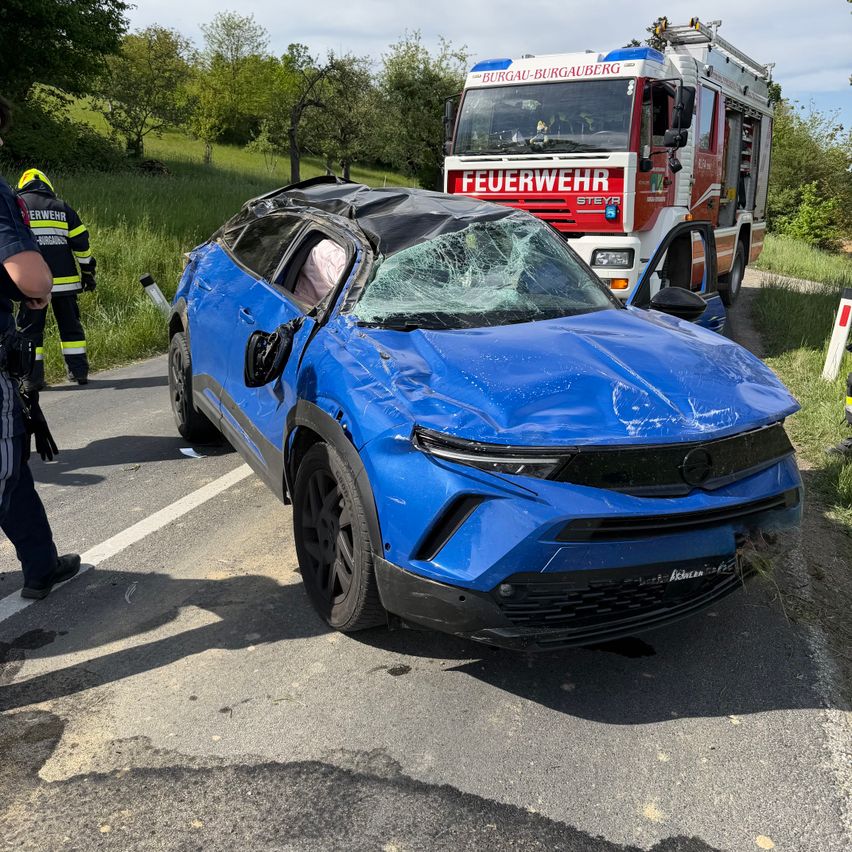 Ein blaues Auto mit eingeschlagener Windschutzscheibe steht am Straßenrand mit einem Feuerwehrfahrzeug dahinter.