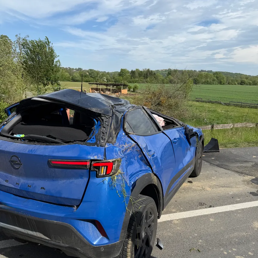 Ein blauer SUV ist in einer ländlichen Umgebung mit einem beschädigten Dach und Stoßfänger. Bäume und ein Zaun sind dahinter, mit einem weitläufigen grünen Feld, das sich in die Ferne erstreckt.