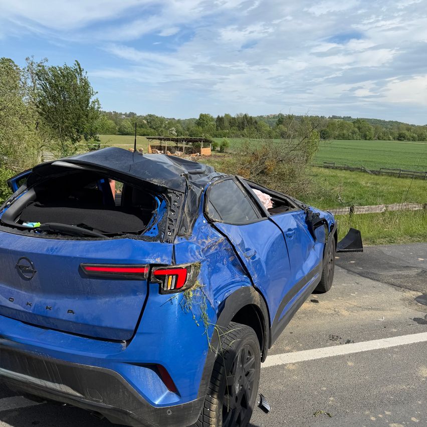 Ein blauer SUV ist in einer ländlichen Umgebung mit einem beschädigten Dach und Stoßfänger. Bäume und ein Zaun sind dahinter, mit einem weitläufigen grünen Feld, das sich in die Ferne erstreckt.