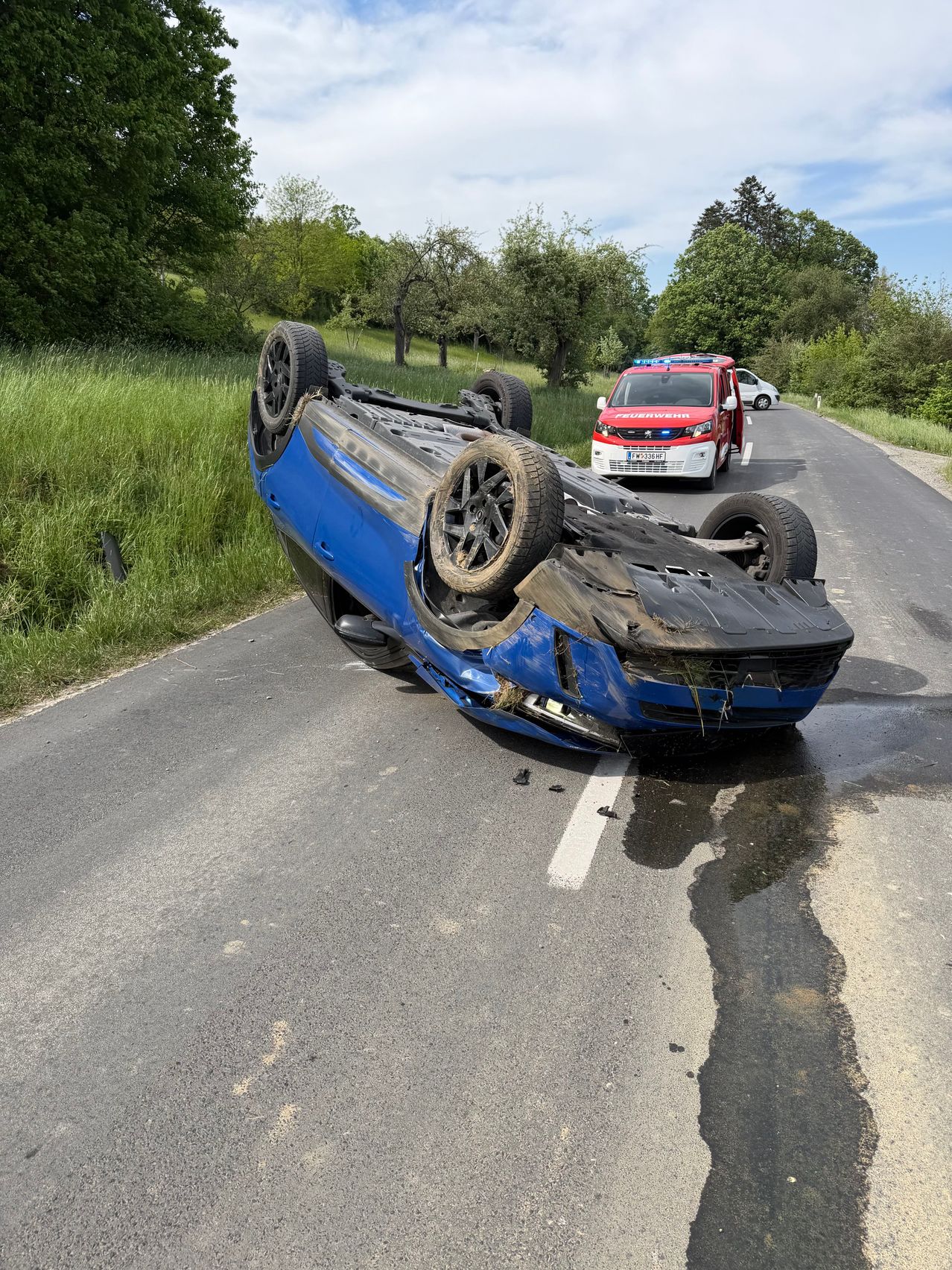 Ein blaues Auto liegt auf der Straße auf dem Kopf, neben Rettungsfahrzeugen. Die Front ist zerquetscht und die Reifen sind abgefallen. Die Straße hat weiße Markierungen und ist von Gras und Bäumen umgeben.