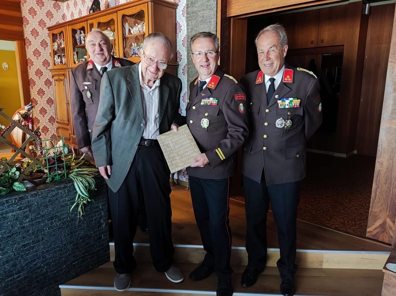 Vier Männer in Uniform stehen auf einer Treppe, einer hält eine gerahmte Geburtstagsnachricht. Der Mann in der Mitte trägt eine Brille und eine Krawatte. Hinter ihnen steht ein hölzerner Schrank mit Glastüren.