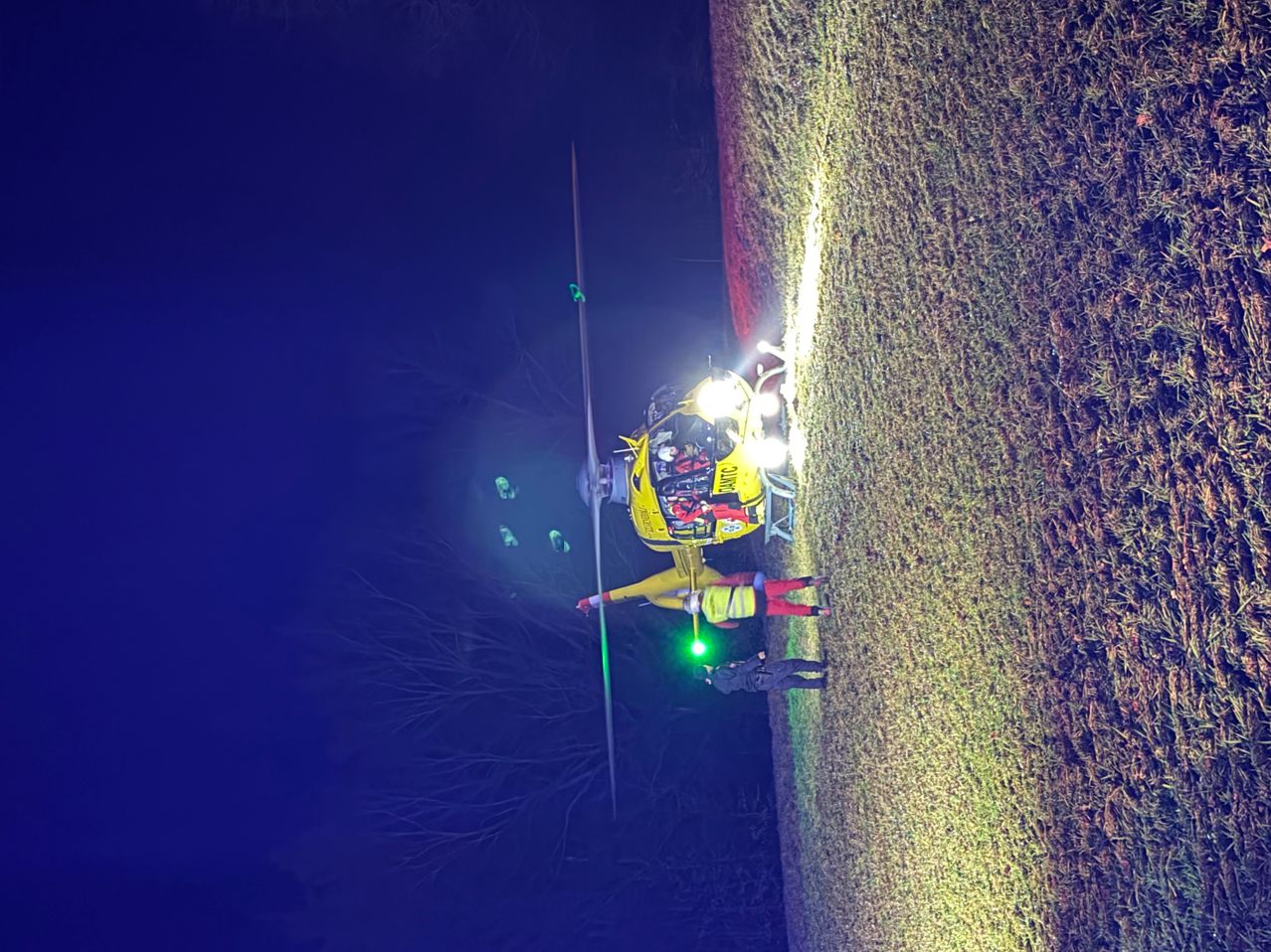 A rescue helicopter is landing on a grassy field at night. Two individuals in yellow safety suits and one in black are on the ground. Green lights are illuminating the scene.