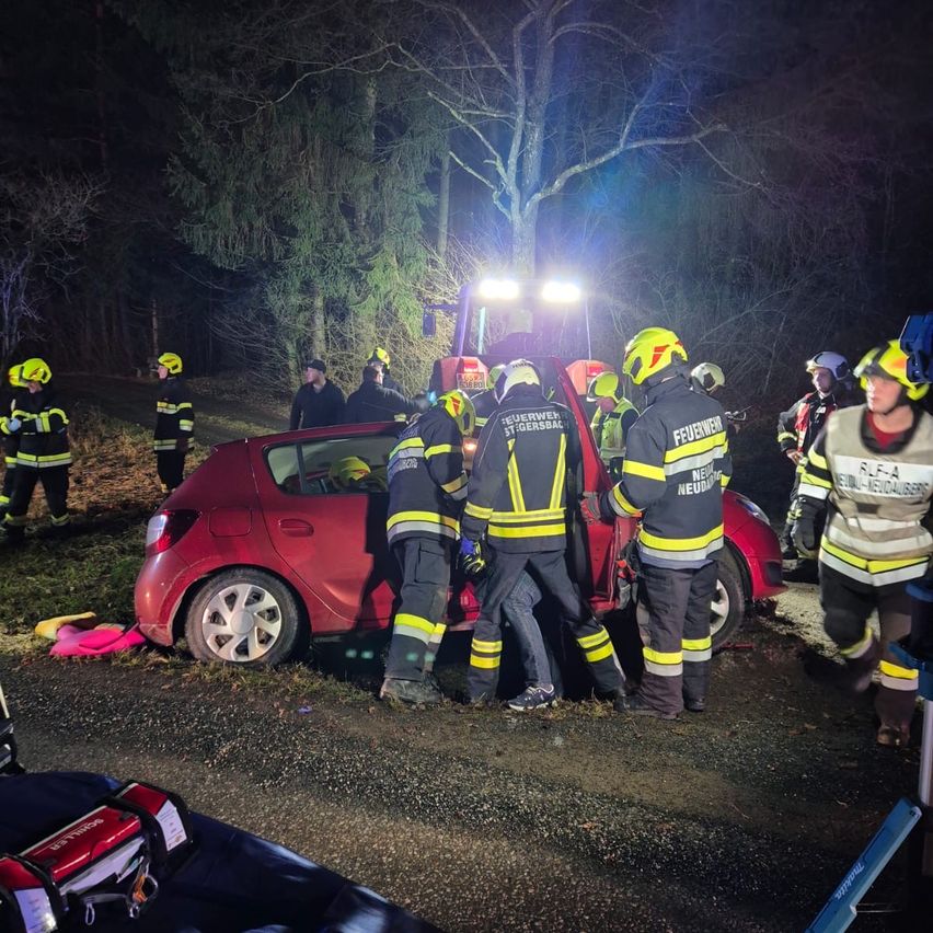 Firefighters are rescuing a person from a crashed red car on a dark road at night. They wear yellow helmets and safety suits.