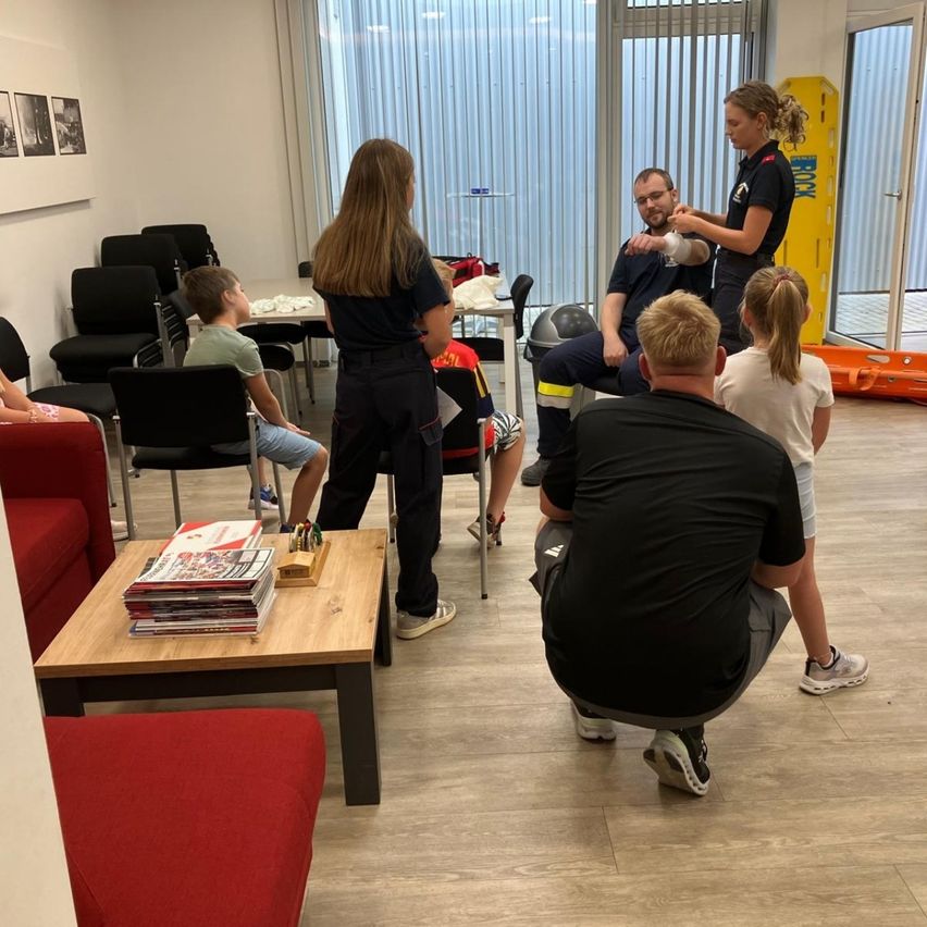 Eine Gruppe von Menschen ist in einem Raum versammelt, wahrscheinlich für eine Schulung. Ein Mann sitzt auf einem Stuhl mit einem Verband am Arm, und eine Frau steht vor ihm. Eine andere Frau steht in der Nähe des Fensters.