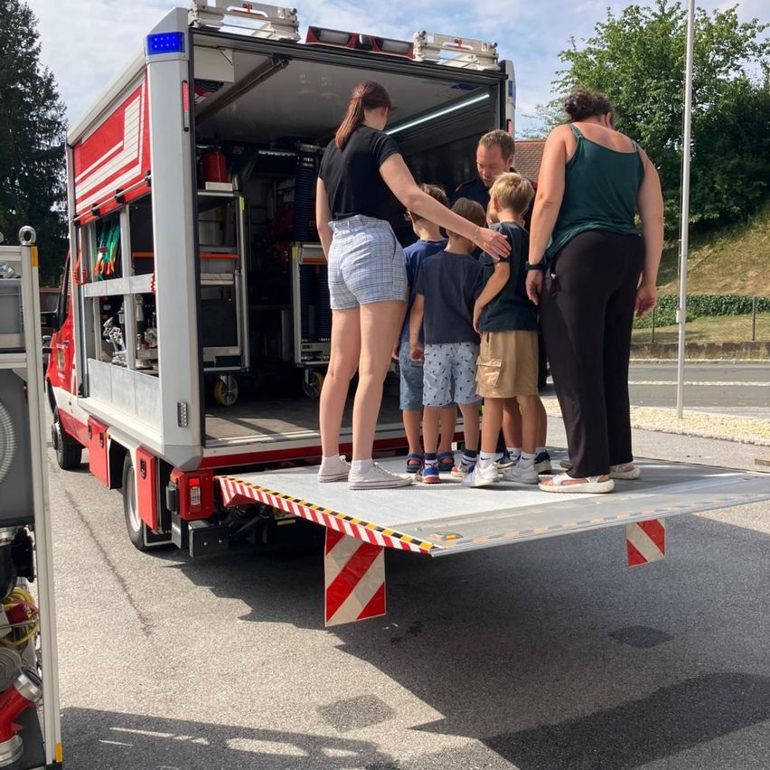 Eine Gruppe von Kindern und Erwachsenen steht auf der Rückseite eines Feuerwehrwagens, wobei die offene Hecktür des Wagens Ausrüstung im Inneren zeigt.