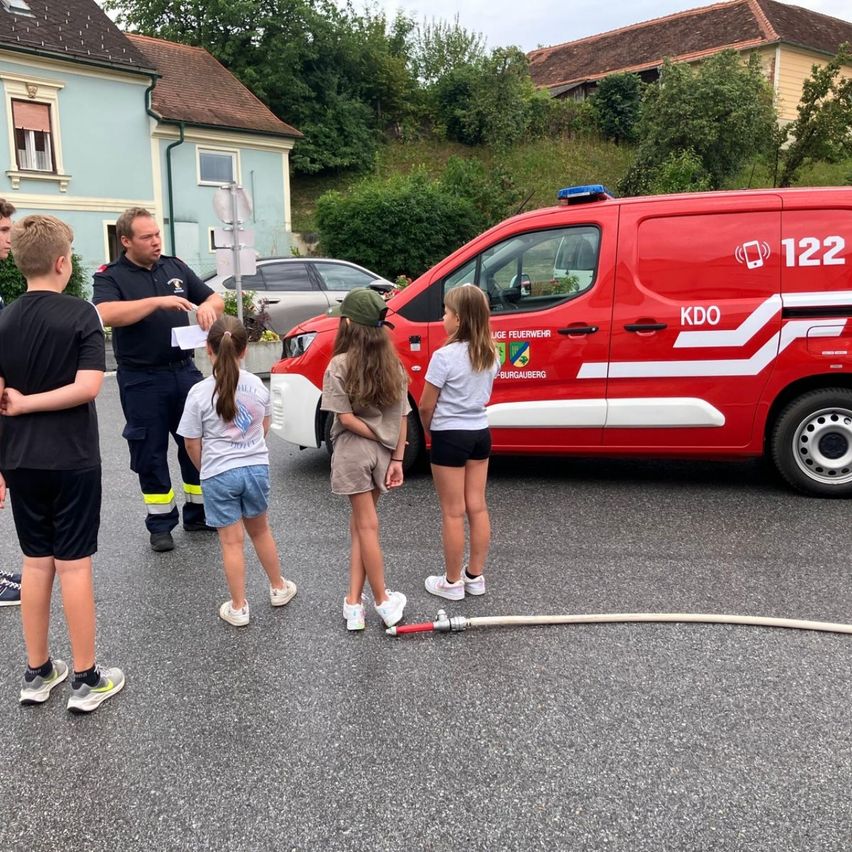 Ein Feuerwehrmann erklärt etwas einer Gruppe von Kindern vor einem roten Feuerwehrwagen.