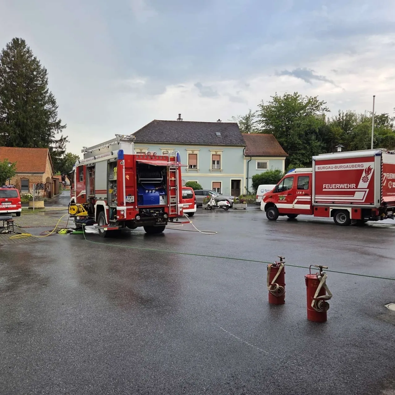Zwei rote Feuerwehrfahrzeuge sind auf einer nassen Straße geparkt, eines mit offener Tür. In der Nähe befinden sich zwei rote Feuerlöscher und ein grüner Schlauch. Hinter den Fahrzeugen stehen Häuser und Bäume.