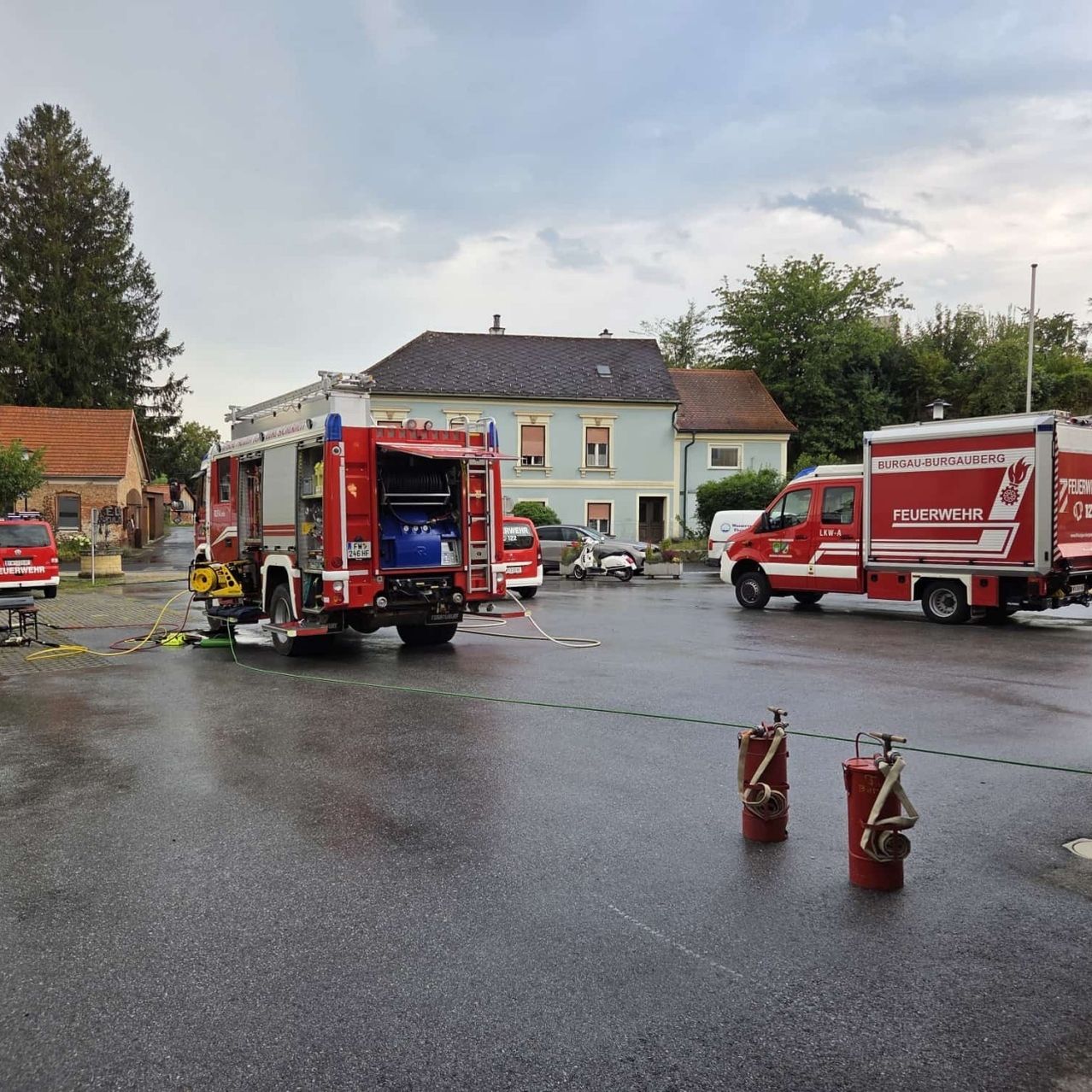 Zwei rote Feuerwehrfahrzeuge sind auf einer nassen Straße geparkt, eines mit offener Tür. In der Nähe befinden sich zwei rote Feuerlöscher und ein grüner Schlauch. Hinter den Fahrzeugen stehen Häuser und Bäume.