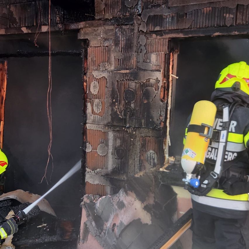 Ein Feuerwehrmann in voller Ausrüstung, einschließlich eines gelben Helms und eines Sauerstofftanks, steht in der Nähe eines verbrannten Gebäudes mit verkohlten Wänden und einer beschädigten Tür. Ein anderer Feuerwehrmann benutzt einen Schlauch, um das Feuer zu löschen.