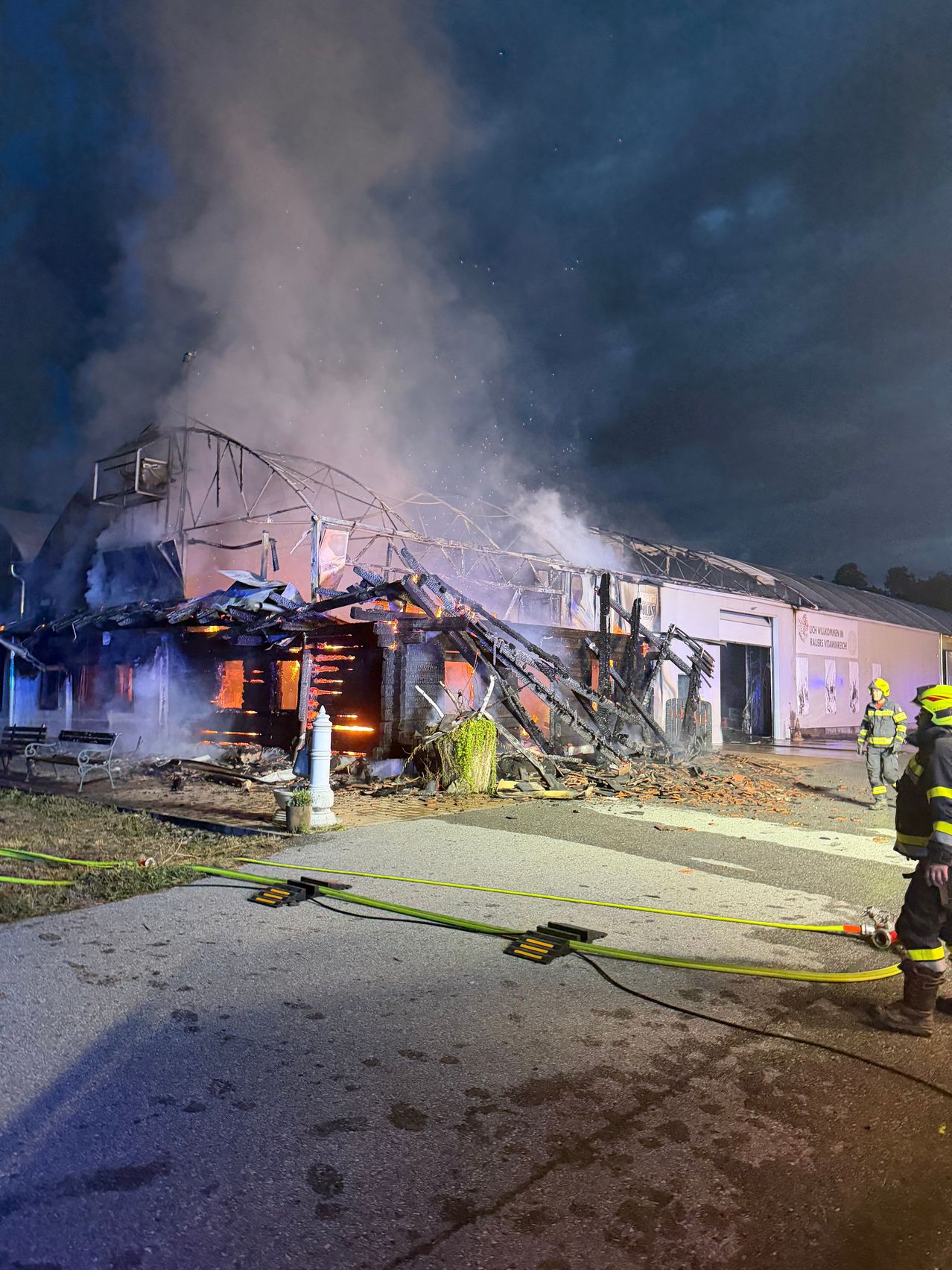 Feuerwehrleute bekämpfen einen Brand in einem Gebäude mit eingestürztem Dach, umgeben von Rauch und Trümmern.