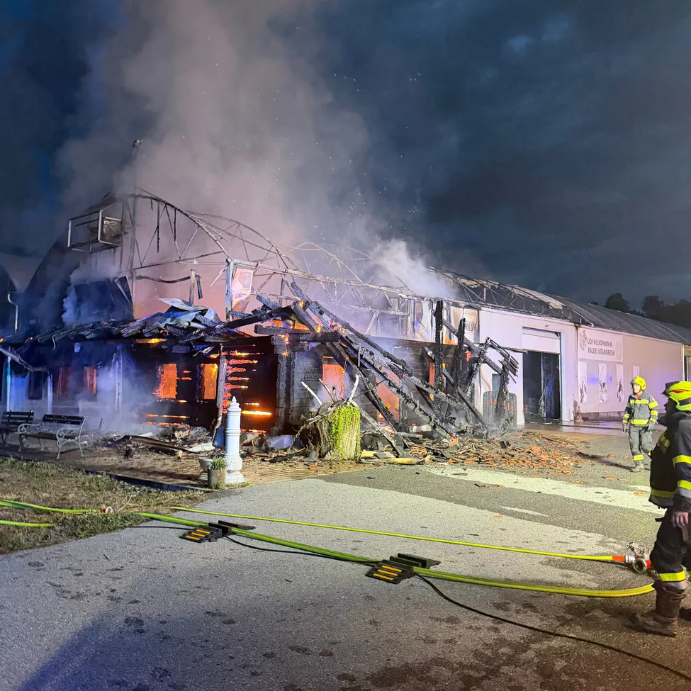 Feuerwehrleute bekämpfen einen Brand in einem Gebäude mit eingestürztem Dach, umgeben von Rauch und Trümmern.