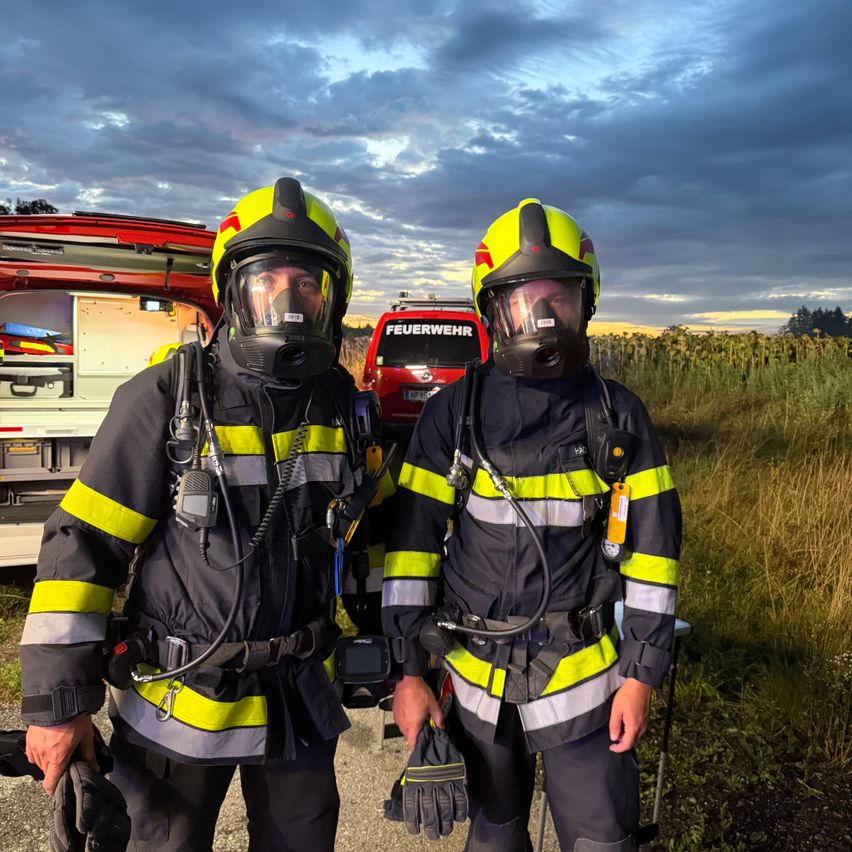 Zwei Feuerwehrleute in voller Ausrüstung stehen neben einem roten Feuerwehrauto mit dem Wort 'Feuerwehr' darauf. Der Himmel ist bewölkt.