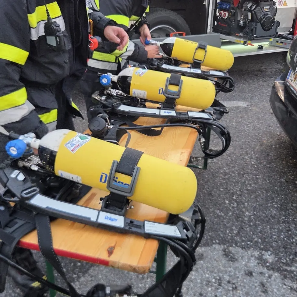 Feuerwehrleute bereiten Sauerstoffflaschen auf einem Holztisch vor. Die Flaschen sind gelb und haben schwarze Gurte. Ein Fahrzeug ist in der Nähe geparkt.