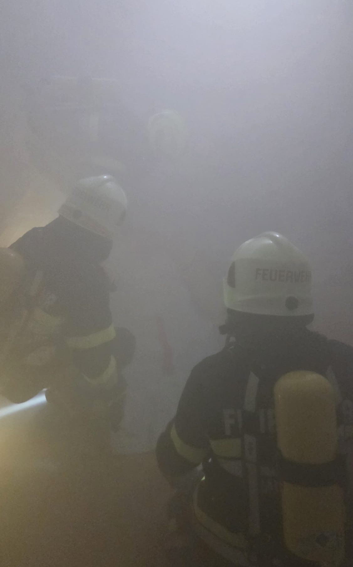 Zwei Feuerwehrleute in Schutzkleidung arbeiten in einer rauchigen Umgebung. Ein Feuerwehrmann hat das Wort 'Feuerwehr' auf seinem Helm.