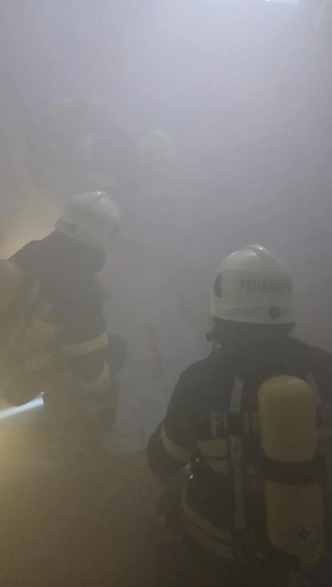Zwei Feuerwehrleute in Schutzkleidung arbeiten in einer rauchigen Umgebung. Ein Feuerwehrmann hat das Wort 'Feuerwehr' auf seinem Helm.