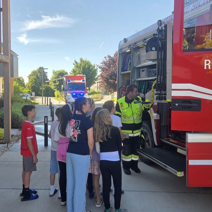 Ein Feuerwehrmann spricht mit Kindern vor einem Feuerwehrwagen auf einem Bürgersteig mit Bäumen und einem Gebäude im Hintergrund.