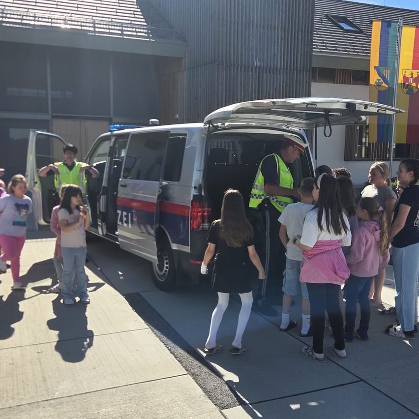 Eine Gruppe von Kindern steht um einen Polizeitransporter herum, dessen Hecktür offen ist. Ein Polizist ist im Inneren des Transporters und spricht mit den Kindern.