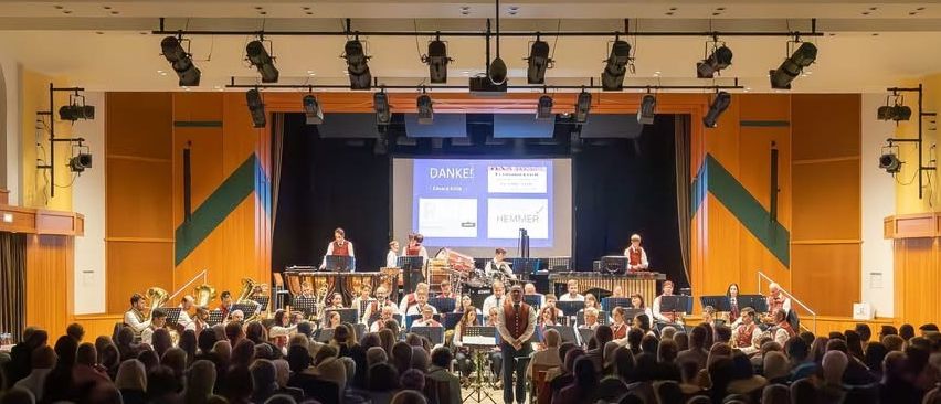 Ein großes Publikum sieht eine Aufführung einer Band in einem Saal. Die Bühne hat Musikinstrumente, eine Leinwand mit 'Danke!' und Scheinwerfer an der Decke.