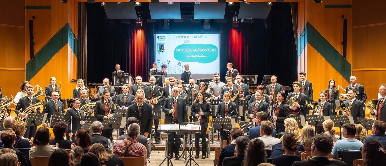 Ein Blechbläserensemble spielt auf einer Bühne vor einem Publikum, mit Scheinwerfern und einer Leinwand im Hintergrund.