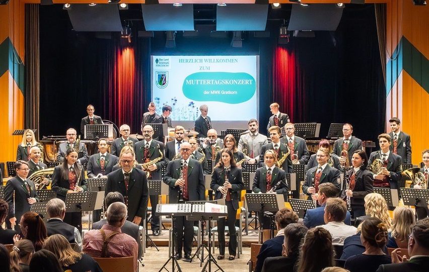 Ein Blechbläserensemble spielt auf einer Bühne vor einem Publikum, mit Scheinwerfern und einer Leinwand im Hintergrund.