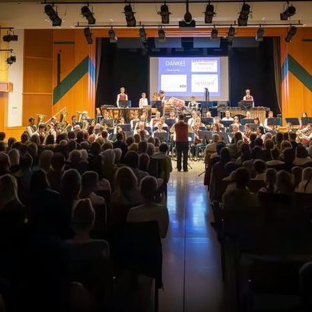 Ein großer Innenbereich mit einer Bühne, auf der eine Band vor einem sitzenden Publikum auftritt. Scheinwerfer sind an der Decke montiert.