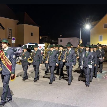 Ein Marschkapelle tritt bei Nacht auf einer Stadtstraße auf, mit Gebäuden auf beiden Seiten und einem Fahrzeug im Hintergrund.