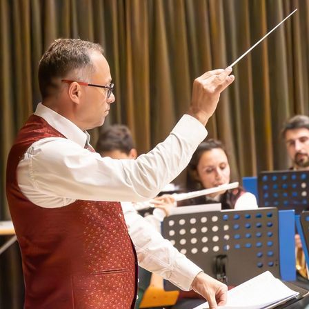 Ein Mann mit Brille dirigiert ein Orchester, mit Musikern, die Flöten und andere Instrumente hinter ihm spielen.