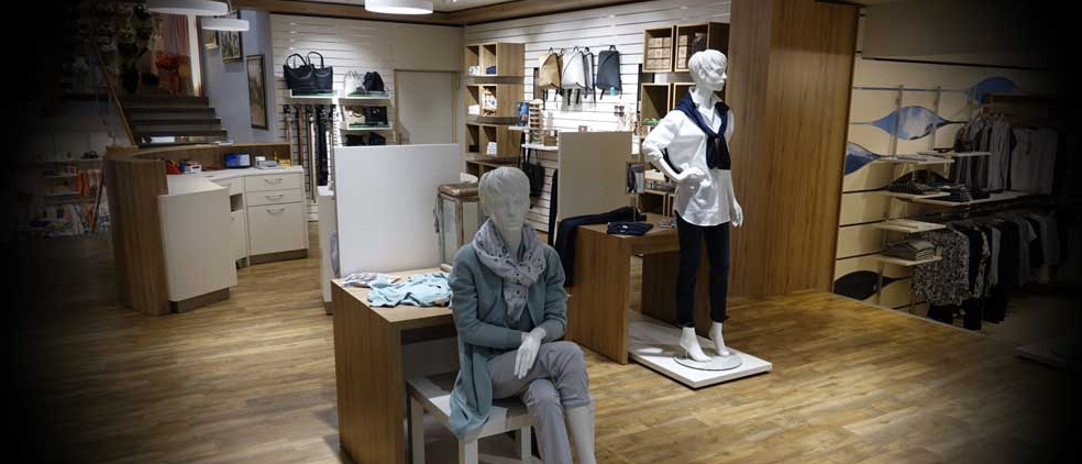 Ein Bekleidungsgeschäft mit Mannequins auf Podesten, die Outfits präsentieren. Holzboden und Regale mit Taschen. Helle Beleuchtung von Deckenlampen.