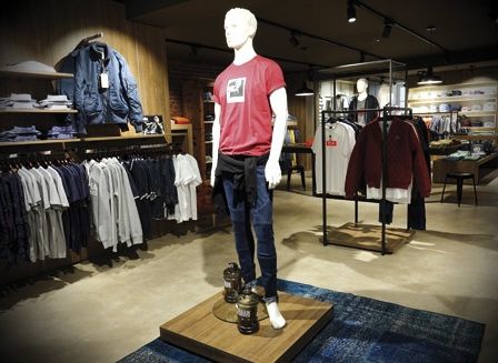 Ein Bekleidungsgeschäft zeigt eine Mannequin in einem roten T-Shirt und Jeans auf einem Holzpodest, umgeben von verschiedenen Hemden an Kleiderständern.
