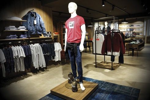 Ein Bekleidungsgeschäft zeigt eine Mannequin in einem roten T-Shirt und Jeans auf einem Holzpodest, umgeben von verschiedenen Hemden an Kleiderständern.
