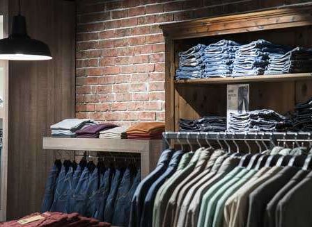 Ein Bekleidungsgeschäft mit einer Ziegelsteinwand zeigt gefaltete Jeans und Hemden auf Regalen und Kleiderbügeln, beleuchtet von einer hängenden Lampe.