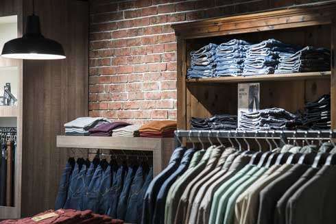 Ein Bekleidungsgeschäft mit einer Ziegelsteinwand zeigt gefaltete Jeans und Hemden auf Regalen und Kleiderbügeln, beleuchtet von einer hängenden Lampe.