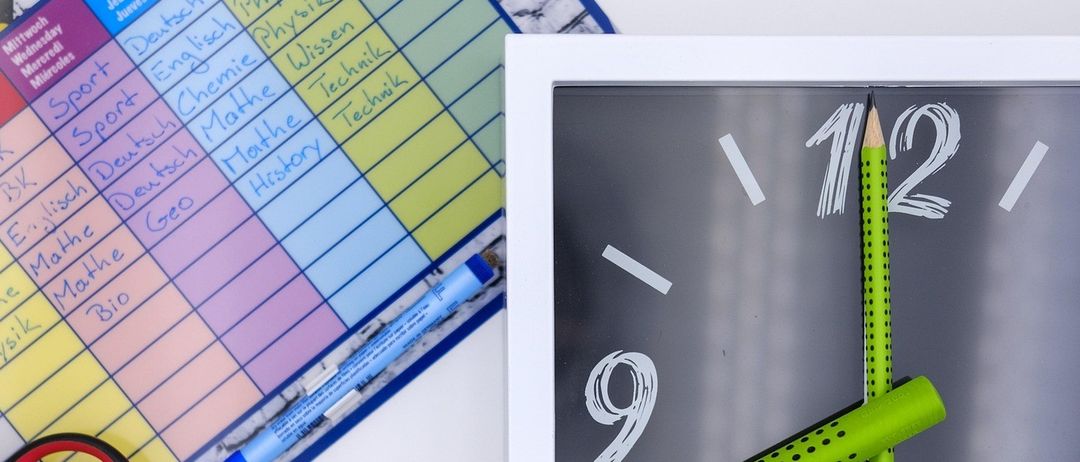 Ein bunter Schulplan mit verschiedenen Fächern ist zu sehen. Daneben befindet sich eine weiß gerahmte Uhr, deren Zeiger auf 10:10 zeigen. Ein Bleistift liegt auf dem Uhrzeiger. Verschiedene farbige Stifte und ein gelber Marker sind in einem Stiftetui.
