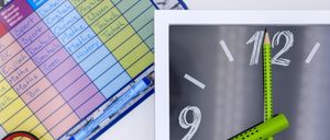 Ein bunter Schulplan mit verschiedenen Fächern ist zu sehen. Daneben befindet sich eine weiß gerahmte Uhr, deren Zeiger auf 10:10 zeigen. Ein Bleistift liegt auf dem Uhrzeiger. Verschiedene farbige Stifte und ein gelber Marker sind in einem Stiftetui.