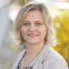 Eine Frau mit kurzen blonden Haaren, blauen Augen und einer Halskette lächelt in die Kamera. Sie trägt einen grauen Blazer und ein gemustertes schwarz-weißes Hemd.