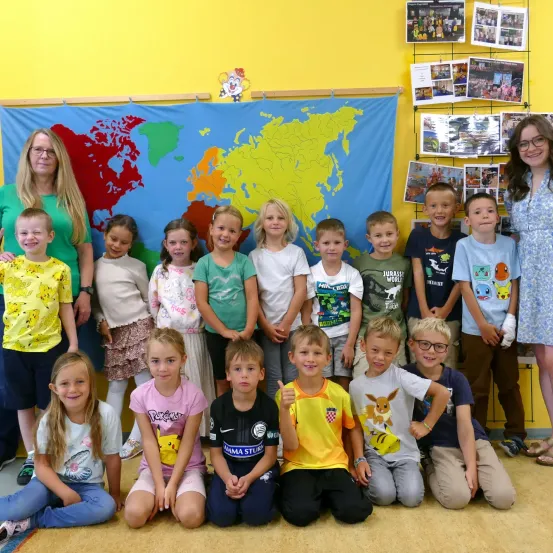 Eine Gruppe von Kindern und zwei Erwachsenen posiert für ein Foto in einem Klassenzimmer mit einer Weltkarte im Hintergrund.