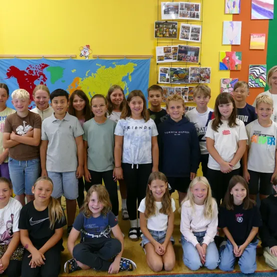 Ein Klassenzimmer mit einer Weltkarte und bunten Postern an der Wand. Schüler in lässiger Kleidung posieren für ein Gruppenfoto vor der Karte.