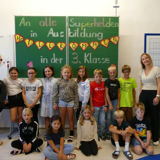 Eine Gruppe von Kindern und ihrer Lehrerin posiert vor einer Tafel mit der Aufschrift 'Willkommen alle Superhelden in Klasse 3'. Die Kinder tragen Freizeitkleidung und einige haben eine Brille. Die Lehrerin lächelt.