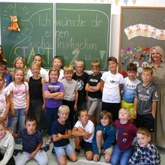 Eine Gruppe von Kindern und einer Frau stehen vor einer Tafel mit einem Fuchs und Text. Die Kinder tragen verschieden farbige Hemden und Hosen. Die Frau lächelt.