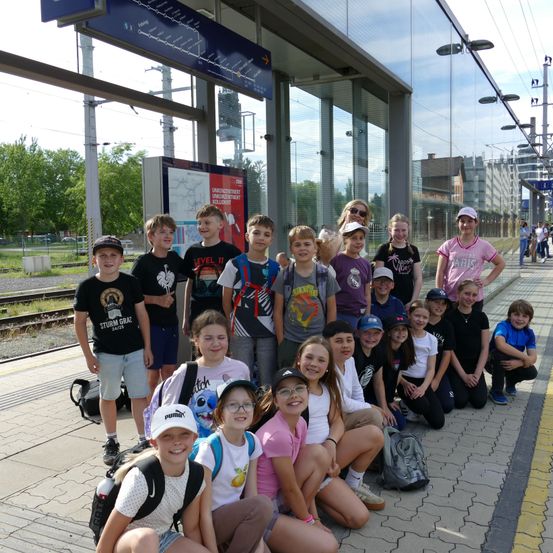 Eine Gruppe von Kindern und Jugendlichen posiert für ein Foto am Bahnhof. Sie lächeln und tragen verschiedene Kleidungsstücke. Der Bahnhof hat eine Glaswand und es gibt Gleise im Hintergrund.