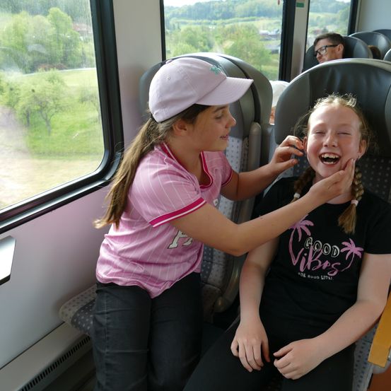 Zwei Mädchen in einem Zug, eines trägt eine rosa Mütze und hilft dem anderen Mädchen mit ihren Haaren, sie lachen beide.