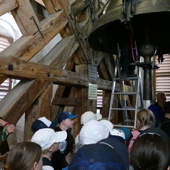 Eine Gruppe von Kindern und Erwachsenen versammelt sich unter einer Holzkonstruktion und beobachtet eine große Glocke. Eine Person steht auf einer Leiter in der Nähe der Glocke.
