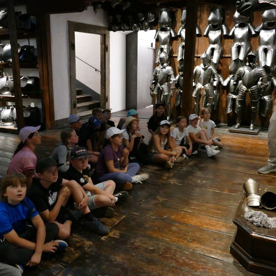 Eine Gruppe von Kindern sitzt auf einem Holzboden und schaut nach oben. Sie befinden sich wahrscheinlich auf einer Museumstour. Dahinter ist Rüstung an der Wand ausgestellt.