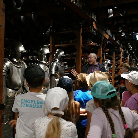 Ein Mann spricht zu einer Gruppe von Kindern in einem Raum voller Rüstungen und Helme zur Ausstellung.