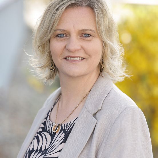 Eine Frau mit kurzen blonden Haaren, die einen hellen Blazer und ein schwarz-weiß gemustertes Oberteil trägt, lächelt und steht draußen.