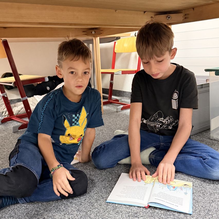 Zwei junge Jungen sitzen auf dem Boden, einer ist in ein Buch vertieft, während der andere zusieht. Hinter ihnen befindet sich eine Klassenzimmerumgebung mit Stühlen und einem Tisch.
