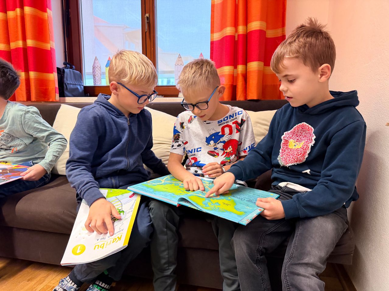 Drei Jungen sitzen auf einer Couch in einem Zimmer und lesen Bücher. Der Junge auf der linken Seite trägt eine Brille und hält ein Buch. Der Junge in der Mitte hat ein Buch auf dem Schoß aufgeschlagen. Der Junge auf der rechten Seite zeigt auf das Buch, das er liest. Hinter ihnen ist ein Fenster mit Vorhängen.