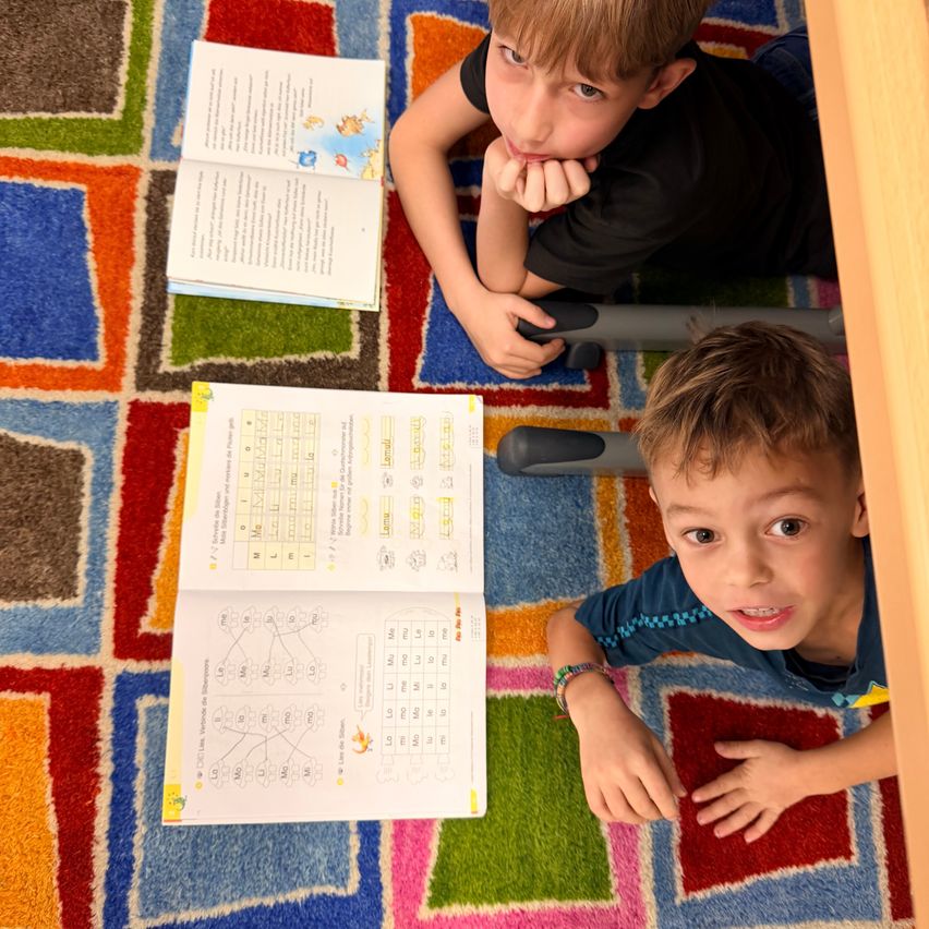 Zwei Jungen liegen auf einem bunten Teppich, einer studiert ein Buch und der andere ein offenes Arbeitsheft mit Rätseln.