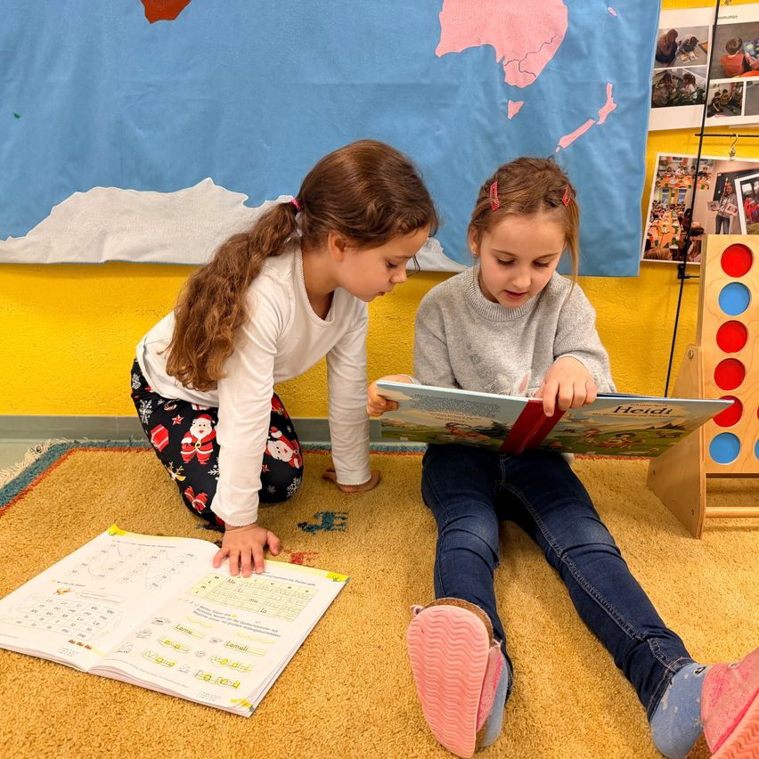 Zwei kleine Mädchen sitzen auf einem Teppich, eine liest ein Buch und die andere schaut in ein offenes Buch mit Zahlen und Symbolen. Der Raum hat eine blaue Wand und einen gelben Boden.