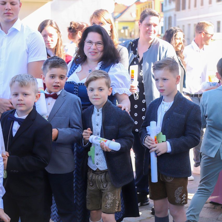 A group of children dressed in traditional Bavarian attire stand together outside. They are holding paper scrolls. Behind them are adults, also dressed in traditional clothing. The setting appears to be a festival.